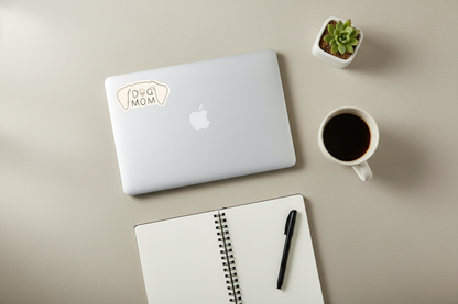 Laptop with 'Mom' sticker, notebook, pen, and coffee cup on a beige surface