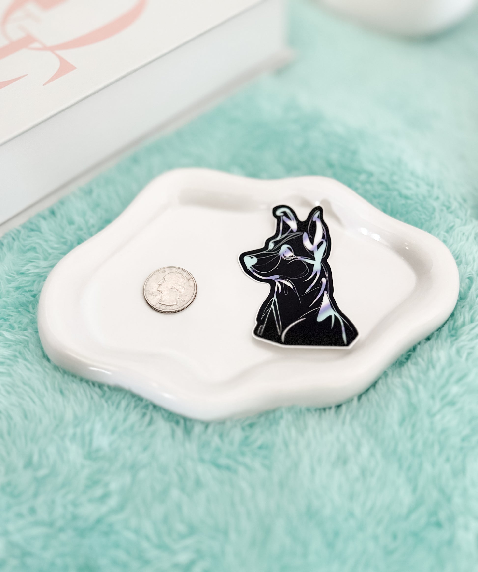 Black dog-shaped pin on a white dish with a coin for scale, on a light blue fabric background